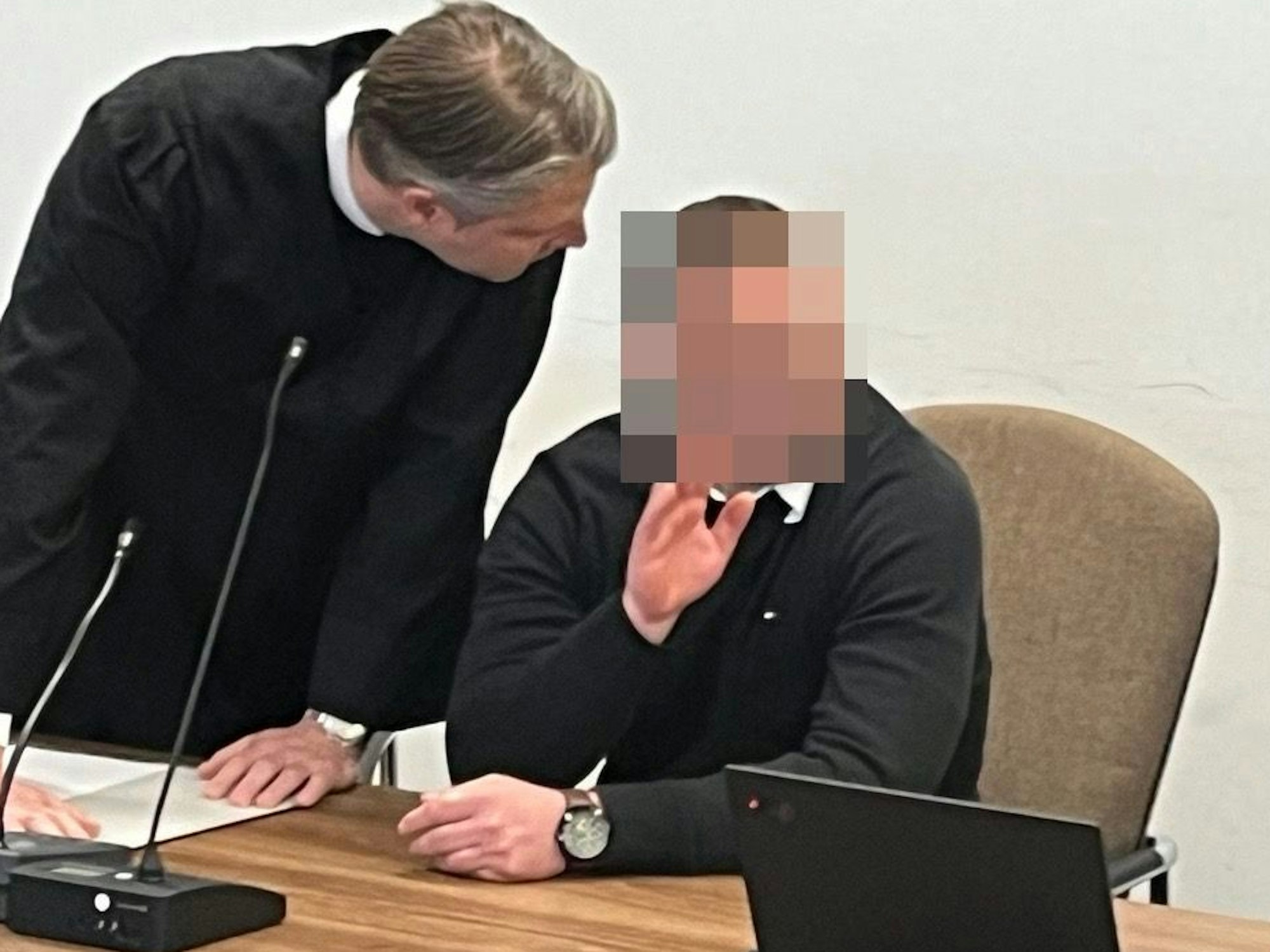 Ein Mann sitzt auf der Anklagebank und winkt, sein Anwalt beugt sich zu ihm runter.