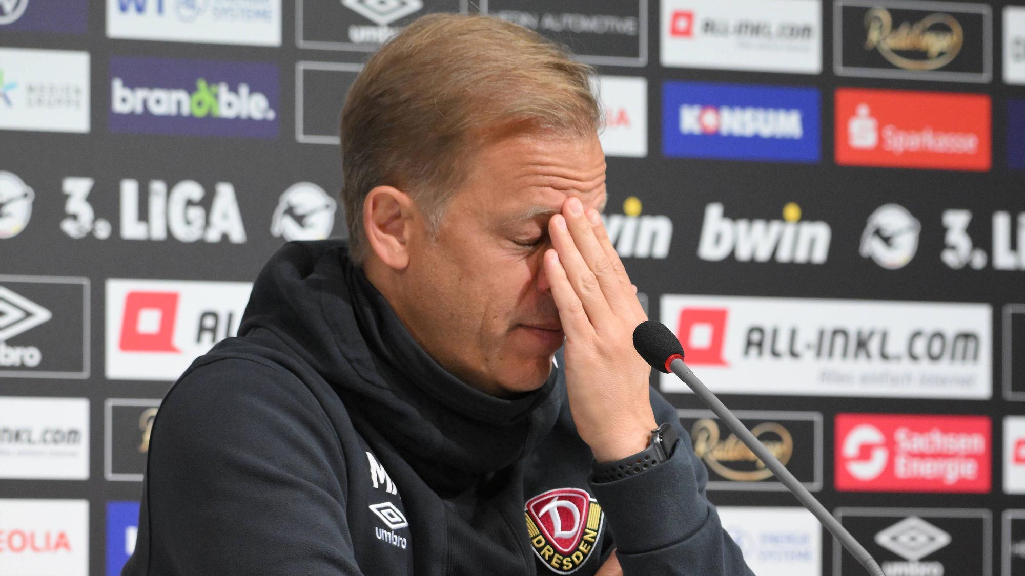 Markus Anfang hält sich bei einer Pressekonferenz von Dynamo Dresden verzweifelt die Hand vor sein Gesicht.