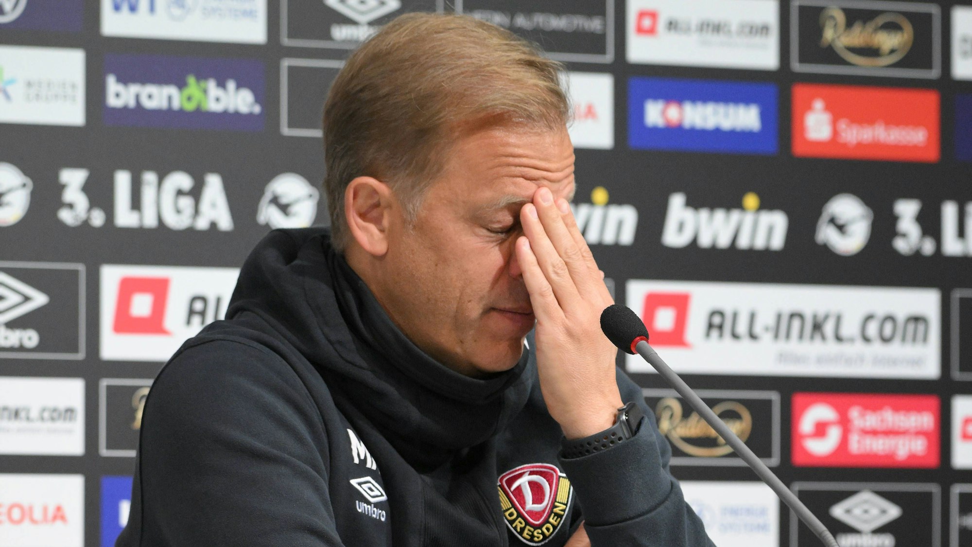 Markus Anfang hält sich bei einer Pressekonferenz von Dynamo Dresden verzweifelt die Hand vor sein Gesicht.