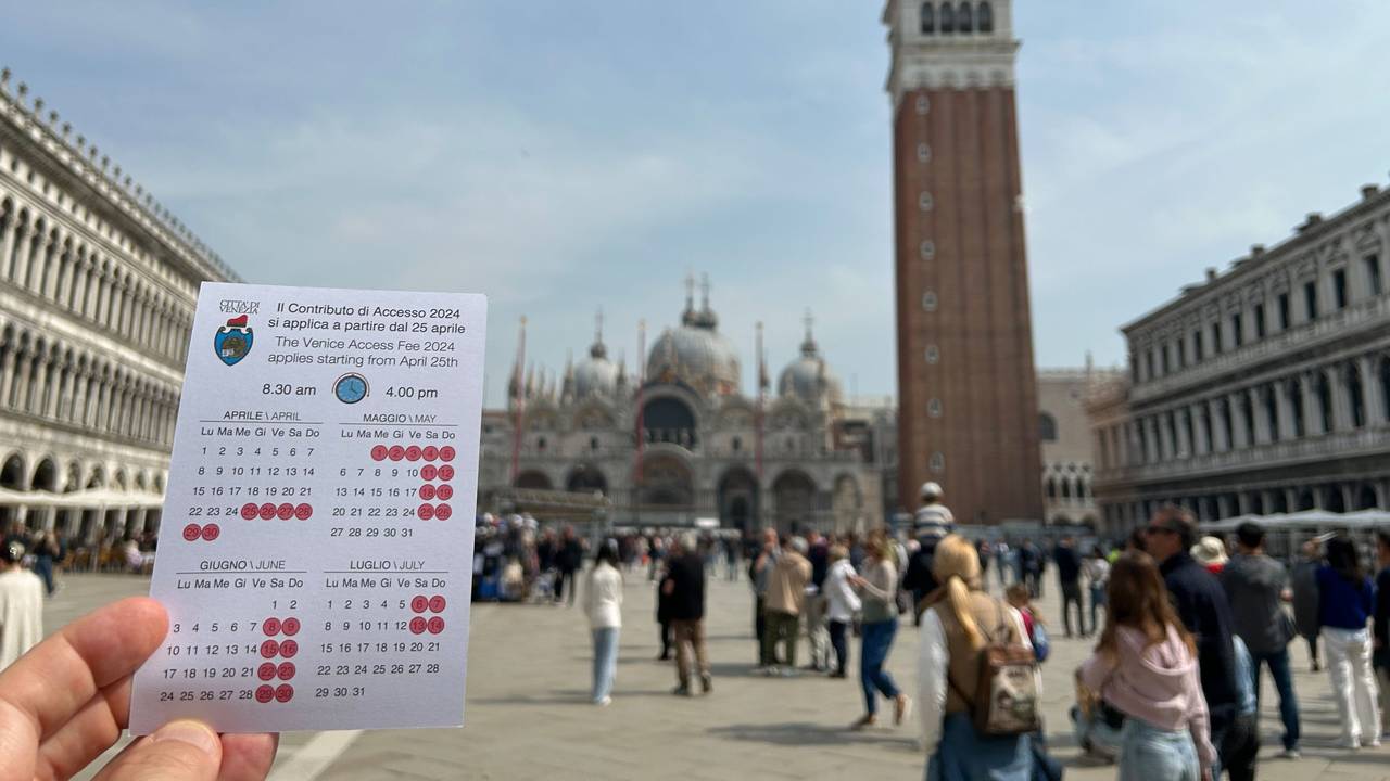 Eine Karte mit dem Kalender für die ersten Tage der Eintrittsgebühr für Venedig, hochgehalten auf dem Markusplatz vor dem Campanile.