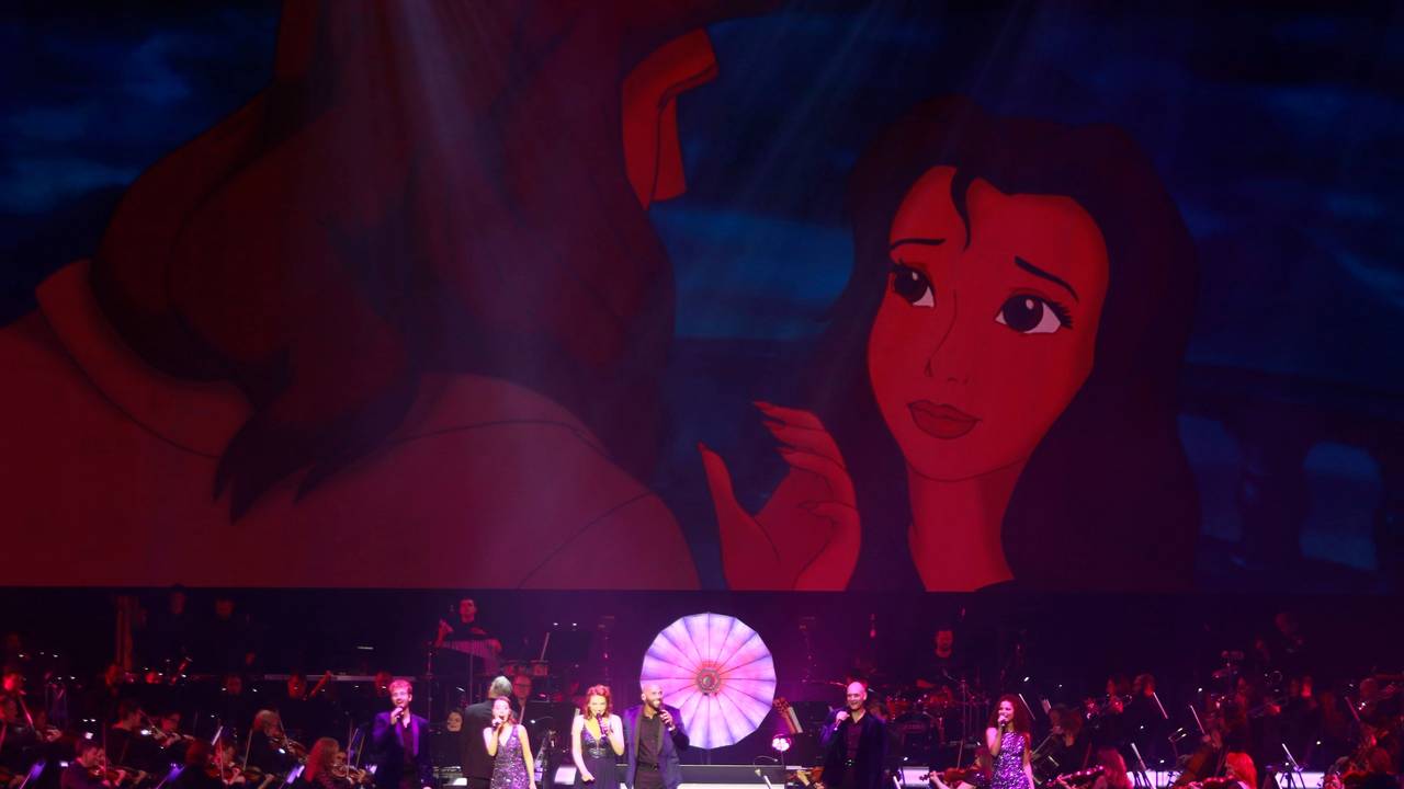 Ein Orchester mit Band und acht Sängern steht auf der Bühne. Im Hintergrund sieht man eine Szene aus Tarzan.