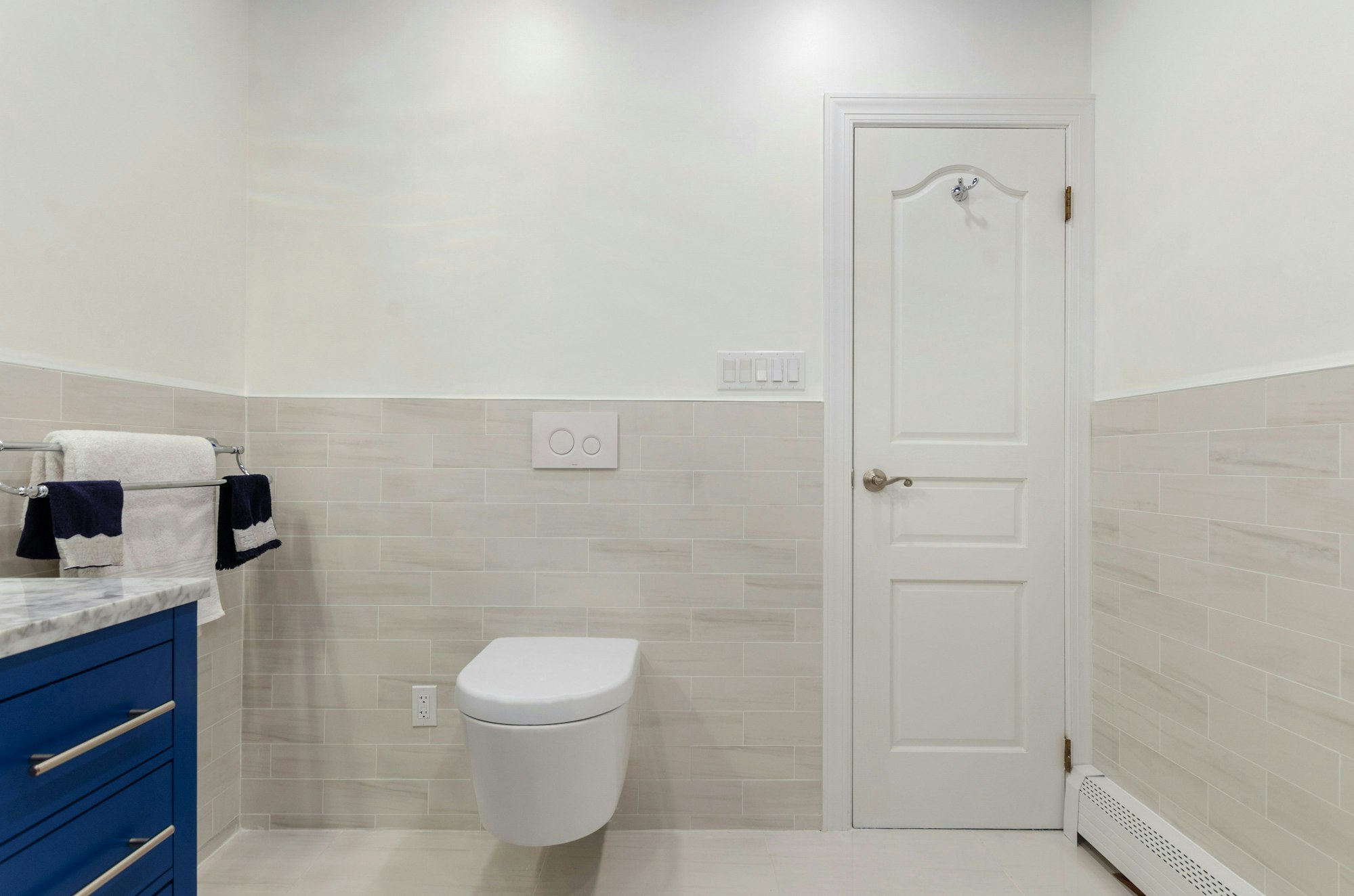 Ein weiß gekacheltes Badezimmer mit einer Toilette in der Bildmitte. Links ist ein Waschbecken, rechts eine Tür.