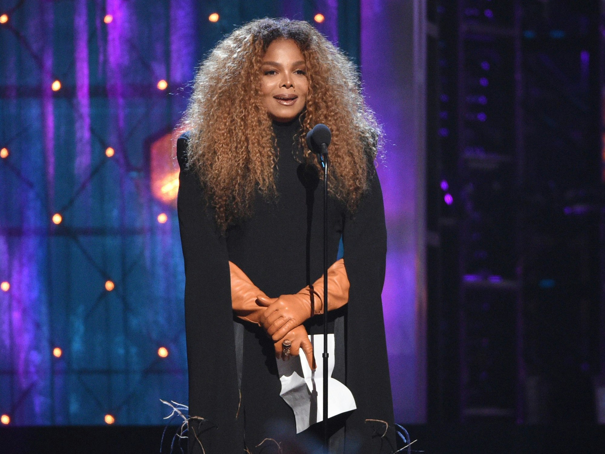 Janet Jackson steht in schwarzem Outfit auf der Bühne und spricht in ein Mikrofon.