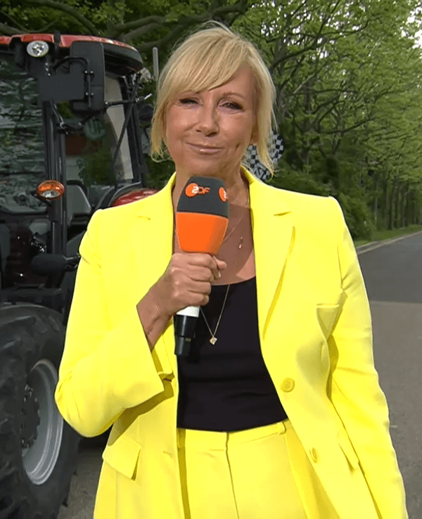 Hier zu sehen: Moderatorin Andrea Kiewel in knallgelbem Anzug, vor einem Traktor bei einer Challenge des ZDF-„Fernsehgarten“ stehend.