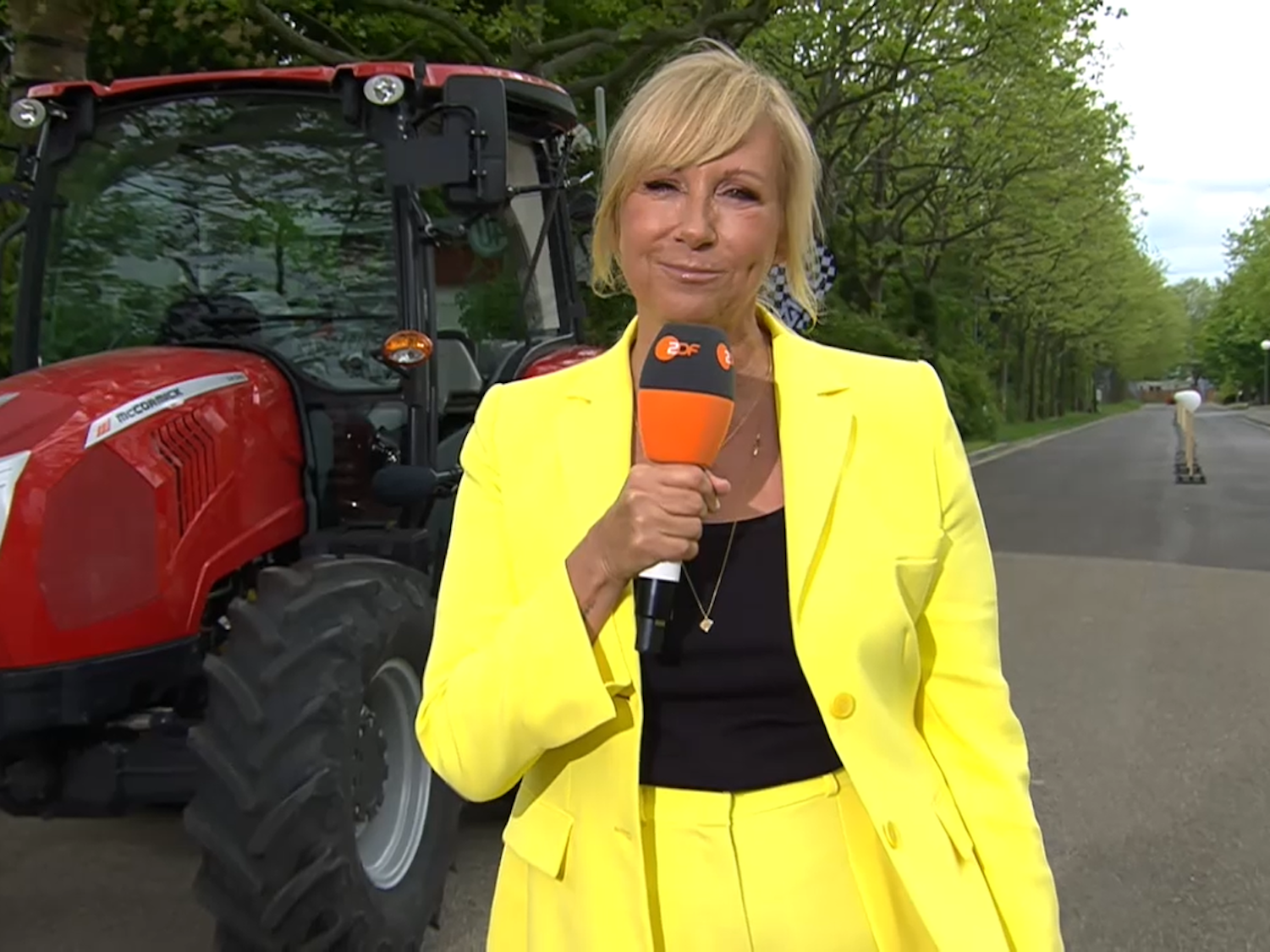 Hier zu sehen: Moderatorin Andrea Kiewel in knallgelbem Anzug, vor einem Traktor bei einer Challenge des ZDF-„Fernsehgarten“ stehend.