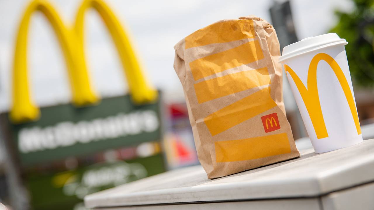 Eine McDonald's To-Go-Tüte und ein To-Go-Becher stehen vor einer McDonald's-Filiale.