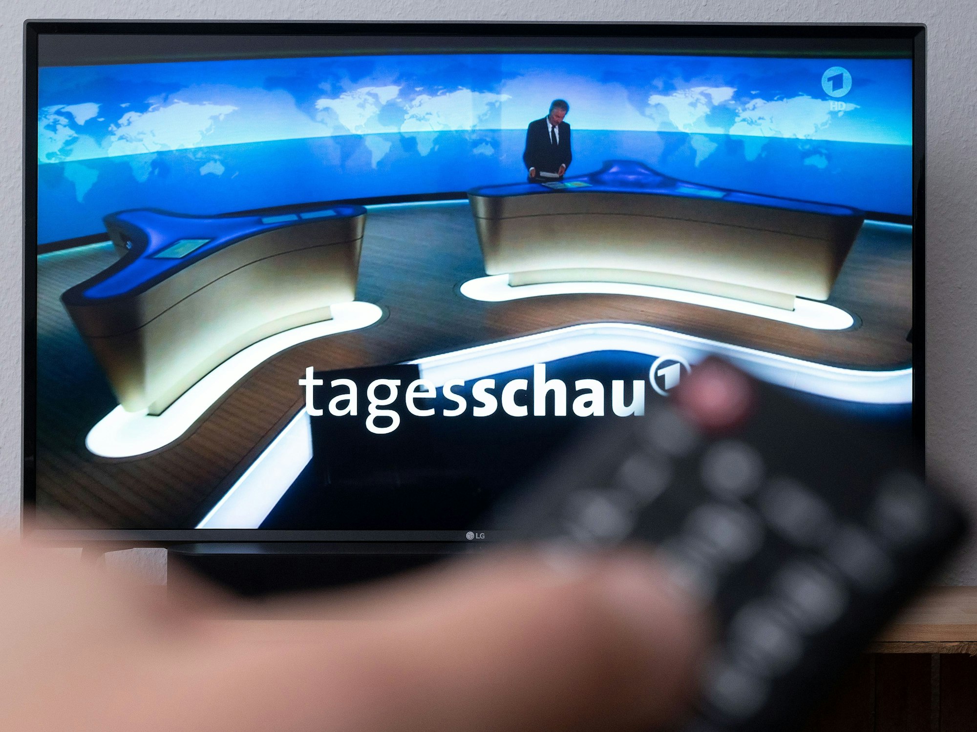 Ein Mann hält eine Fernbedienung vor einen Fernseher, auf dem die „Tagesschau“ läuft.