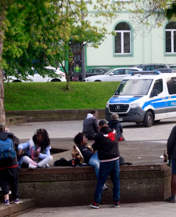 Mehrere Personen sitzen oder liegen auf Steinbänken, andere stehen davor, teilweise haben sie eine Bierflasche dabei.
