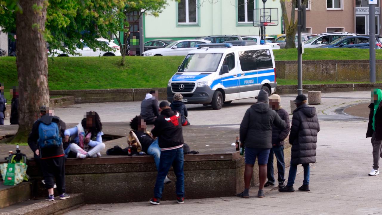 Mehrere Personen sitzen oder liegen auf Steinbänken, andere stehen davor, teilweise haben sie eine Bierflasche dabei.