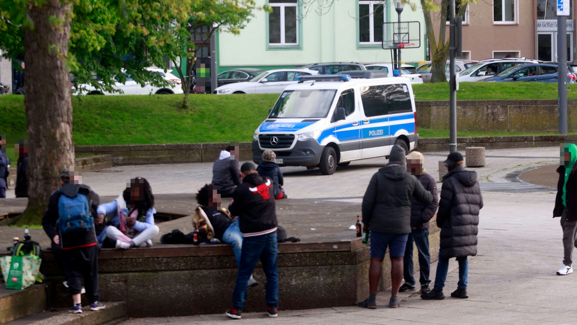 Menschen sitzen an einem Platz, im Hintergrund steht ein Polizeiwagen.
