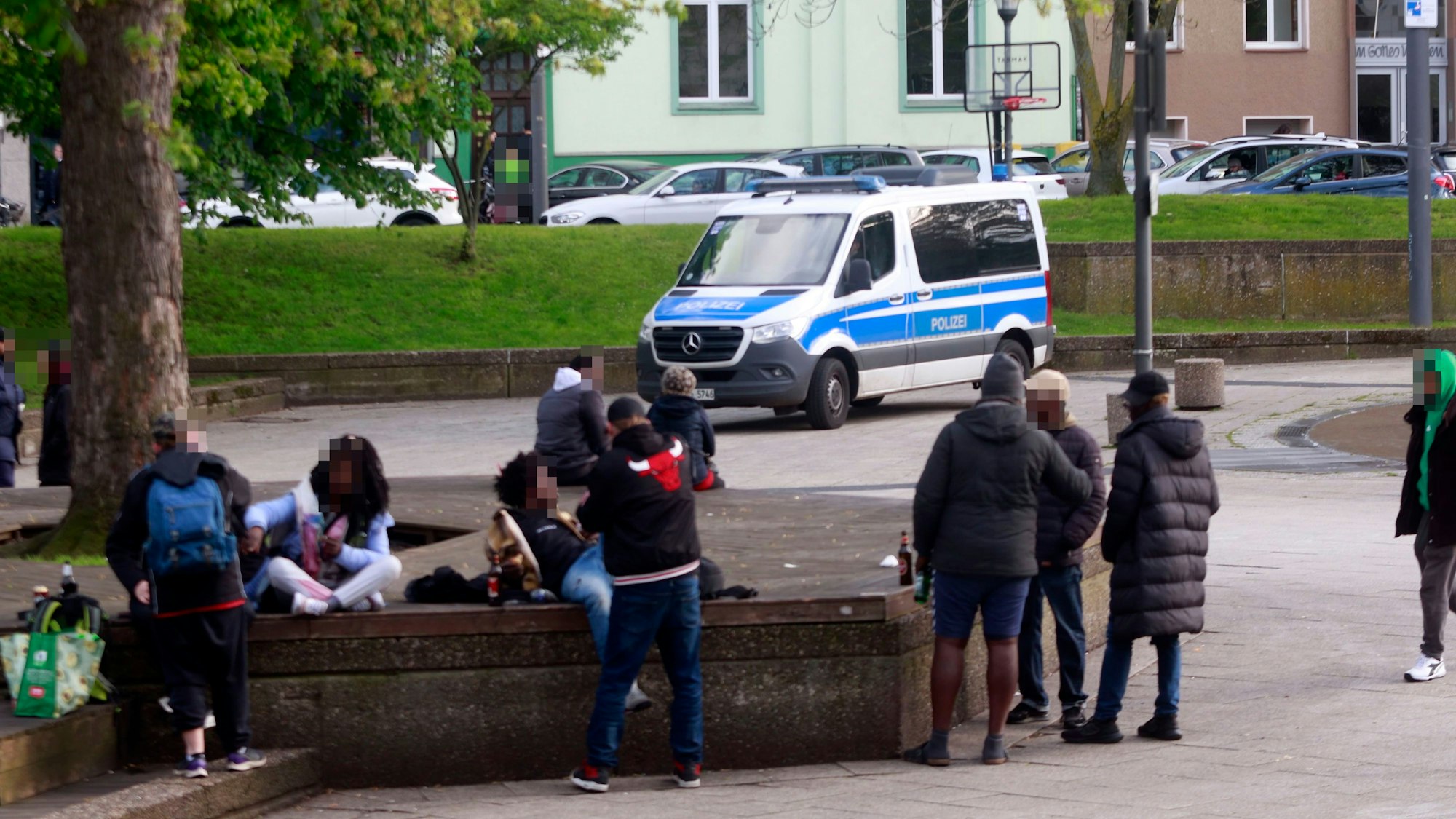 Mehrere Personen sitzen oder liegen auf Steinbänken, andere stehen davor, teilweise haben sie eine Bierflasche dabei.