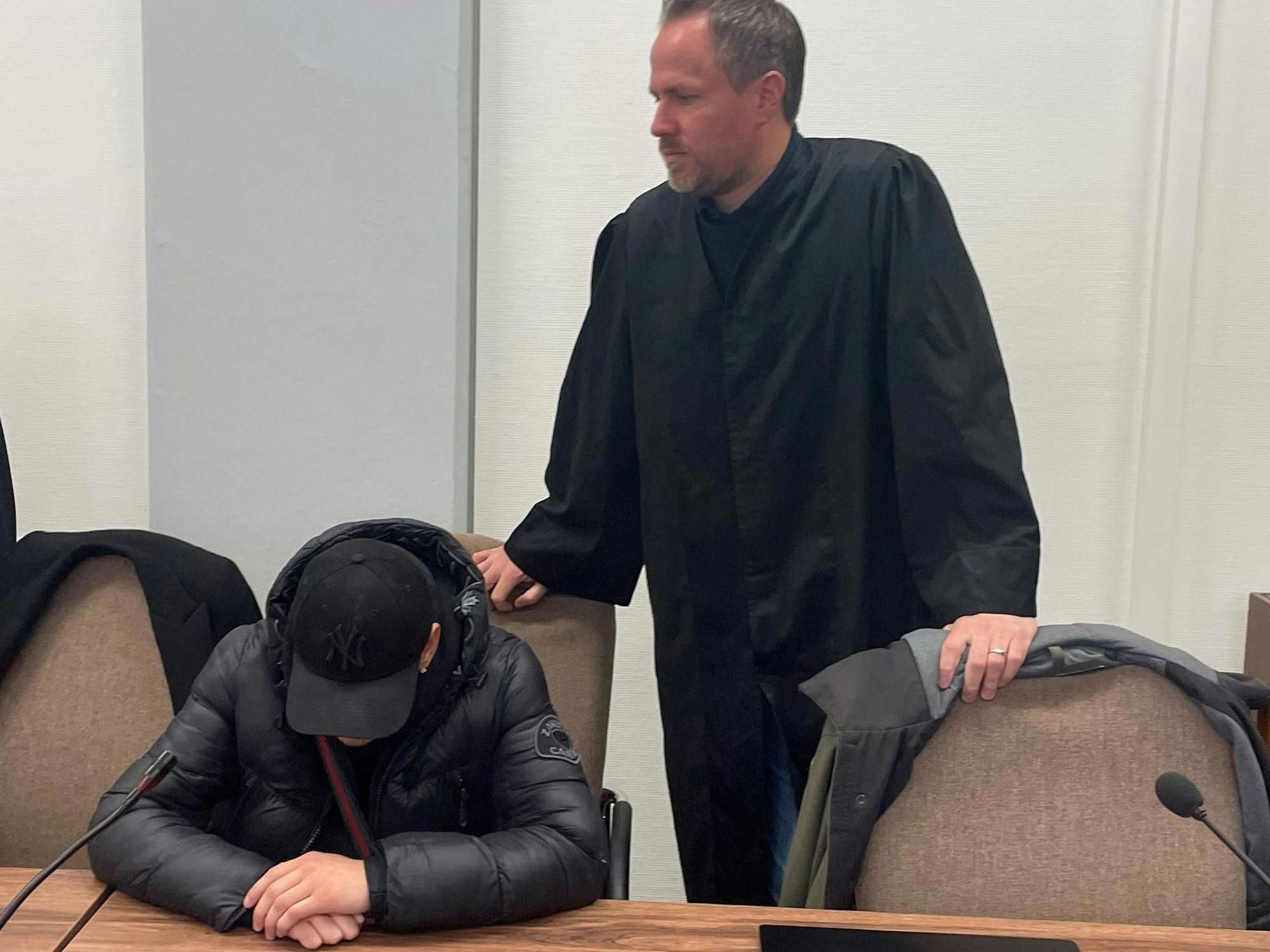 Ein junger Mann sitzt mit gesenktem Kopf auf der Anklagebank, sein Anwalt steht neben ihm.