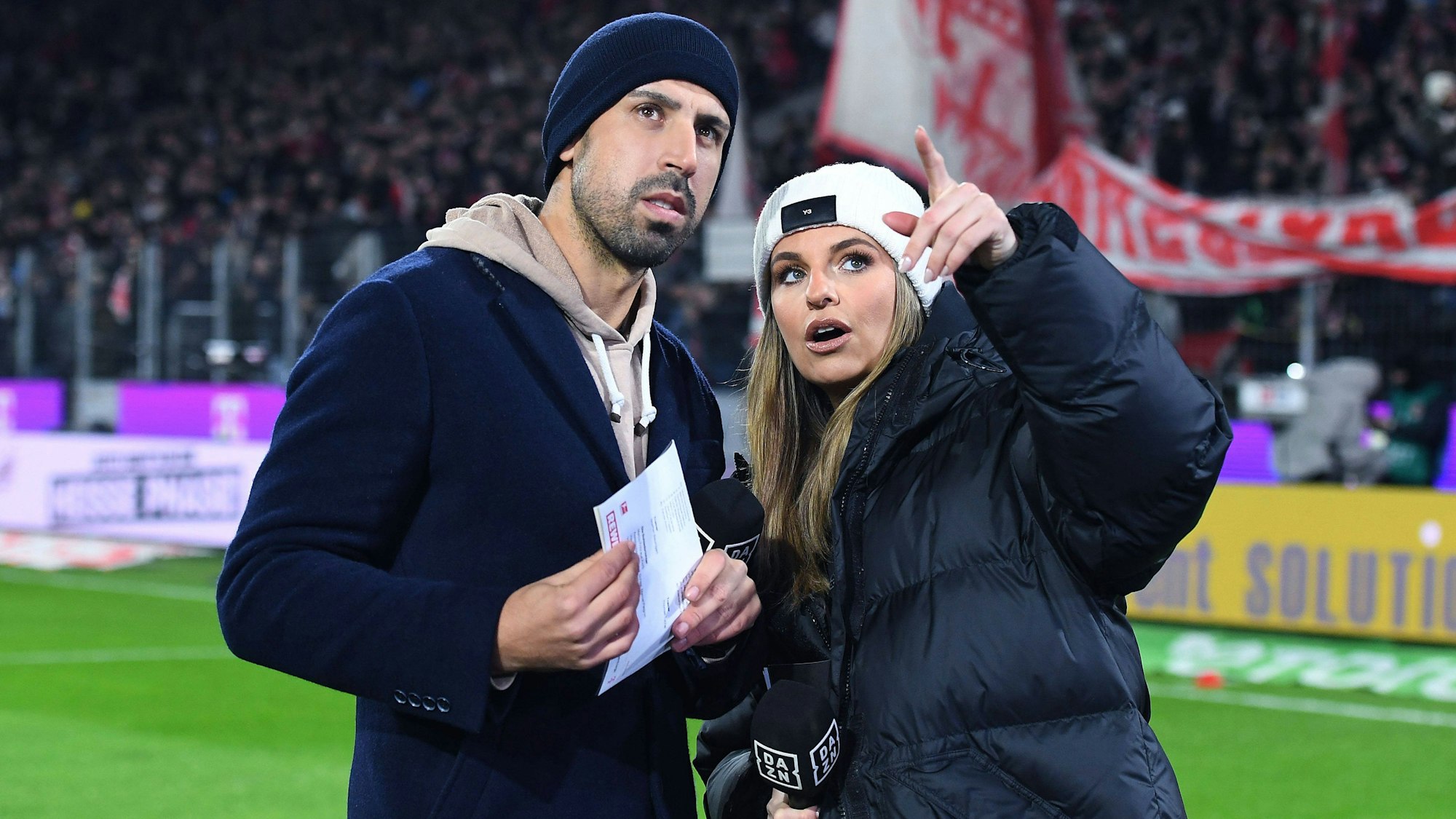 Sami Khedira und Moderatorin Laura Wontorra blicken bei einer DAZN-Übertragung auf die Tribüne im Stadion.