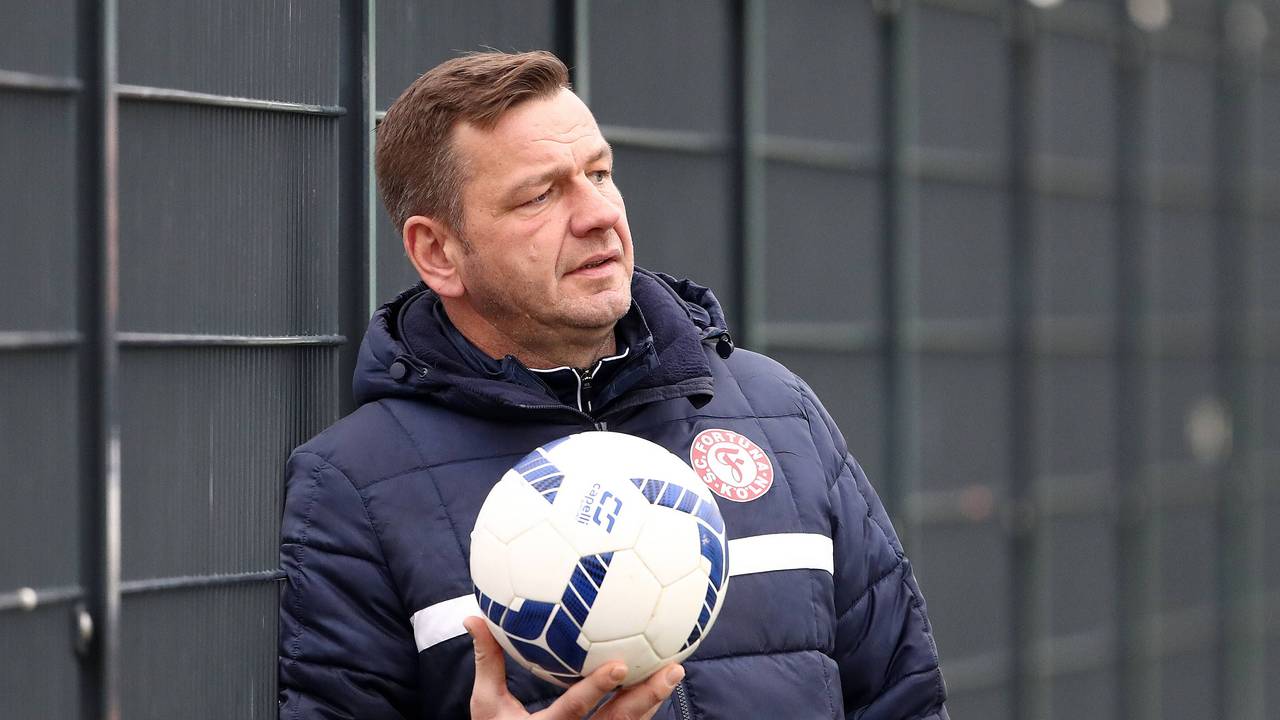 Torwarttrainer Georg Koch lehnt am Zaun und hält einen Ball in der Hand.