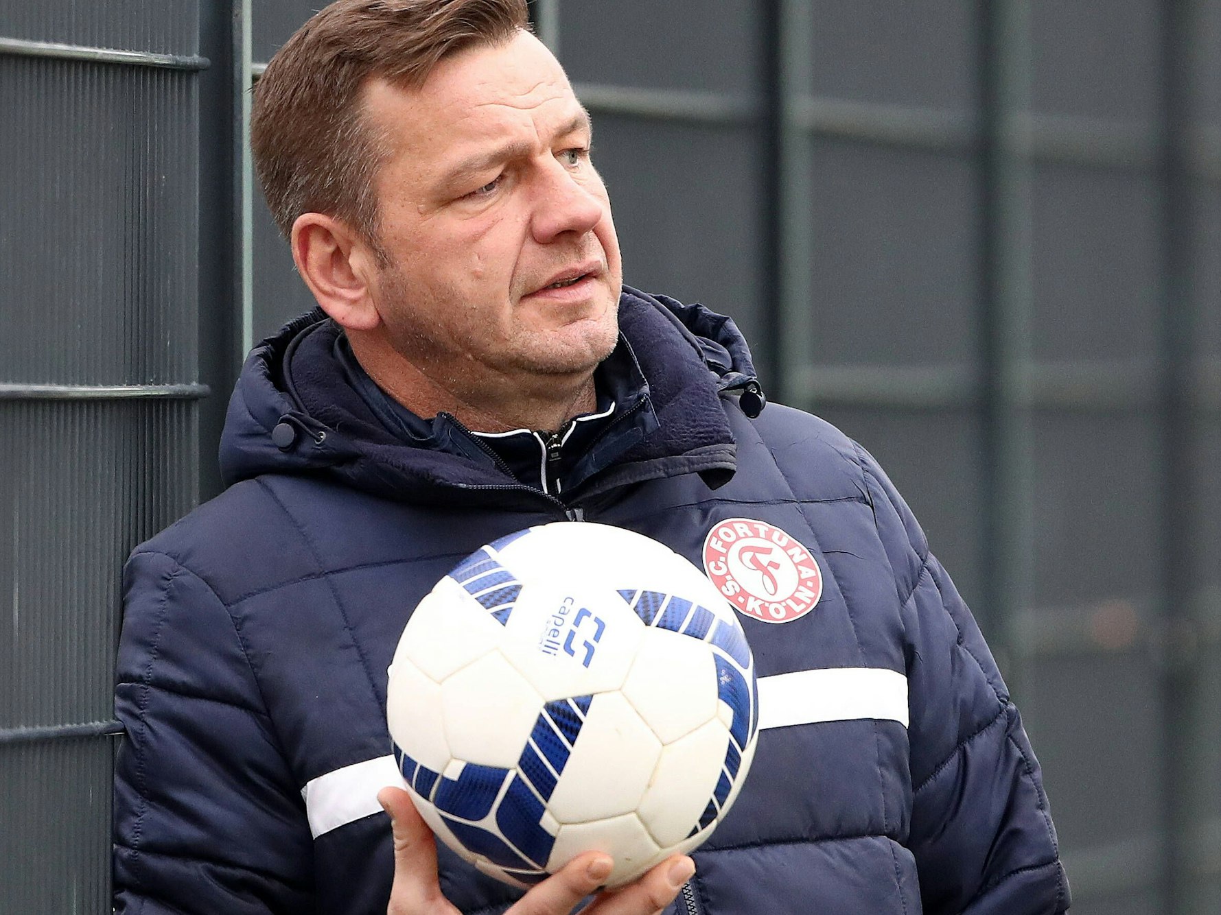 Torwarttrainer Georg Koch lehnt am Zaun und hält einen Ball in der Hand.