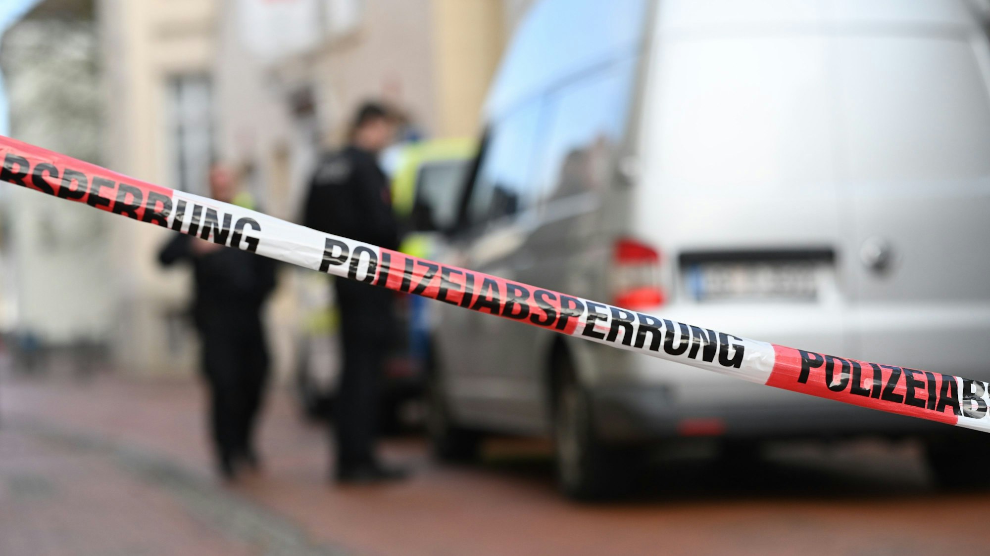 Eine Straße ist mit Flatterband abgesperrt, dahinter stehen zwei Fahrzeuge sowie zwei Polizisten.