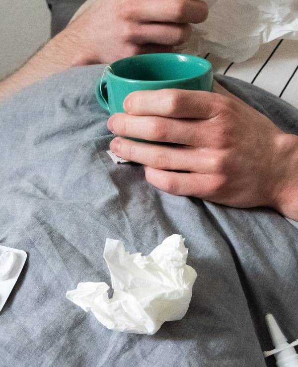Ein Mann liegt mit Taschentüchern, Teetasse, Nasenspray und Tabletten im Bett (gestellte Szene).