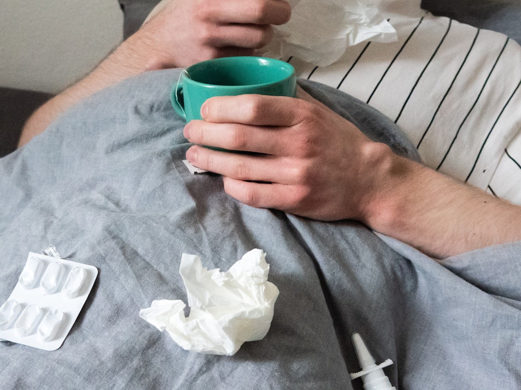 Ein Mann liegt mit Taschentüchern, Teetasse, Nasenspray und Tabletten im Bett (gestellte Szene).
