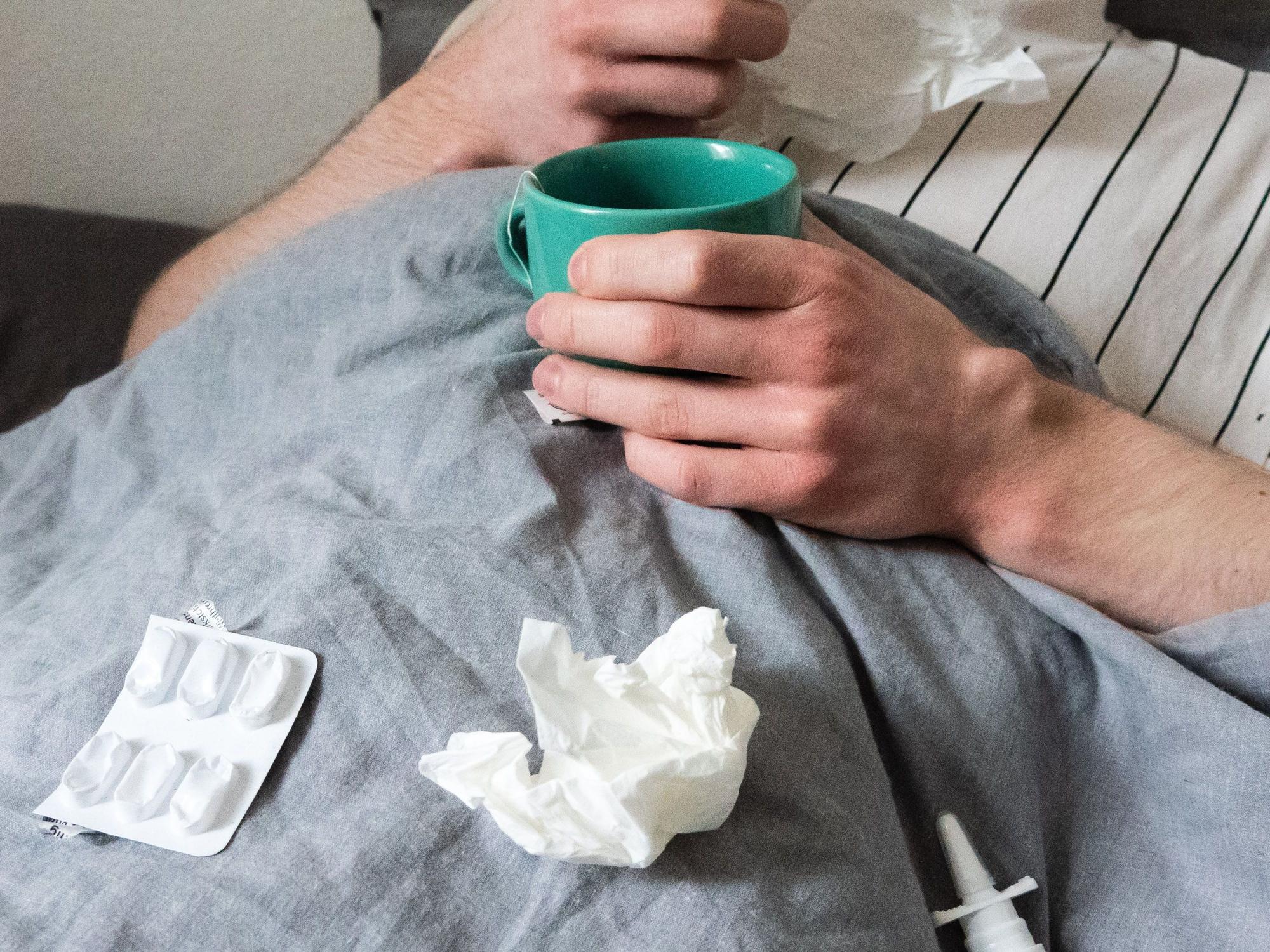 Ein Mann liegt mit Taschentüchern, Teetasse, Nasenspray und Tabletten im Bett (gestellte Szene).