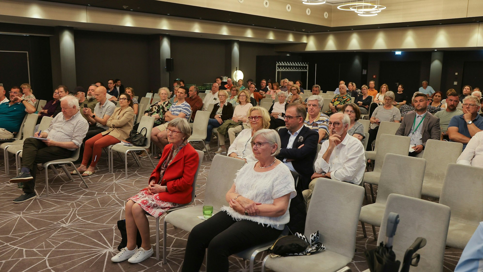 Menschen sitzen auf Stühlen in einem Saal und hören einem Vortrag zu.