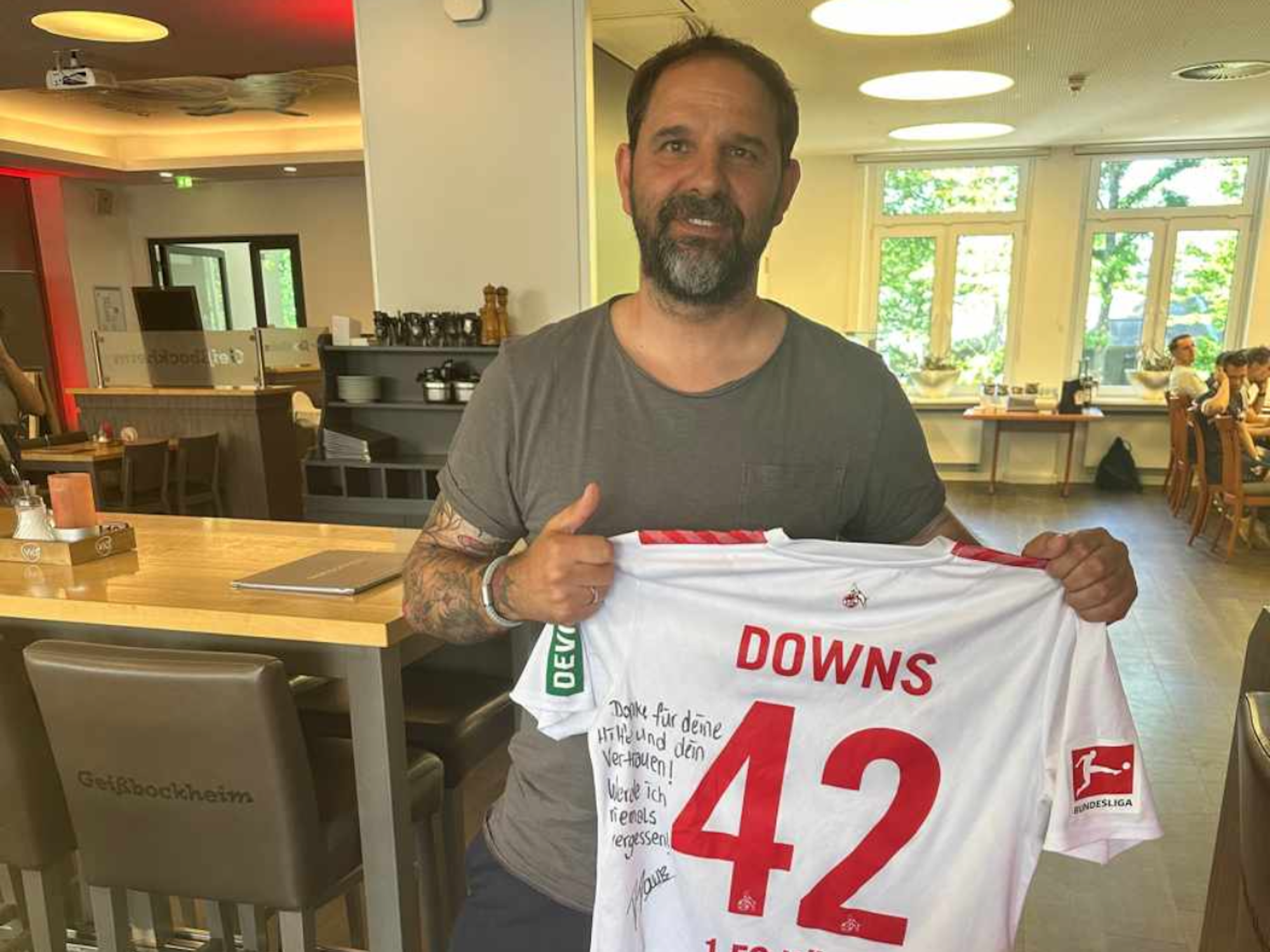 Stefan Ruthenbeck posiert am Geißbockheim mit dem Trikot inklusive Widmung von Damion Downs.
