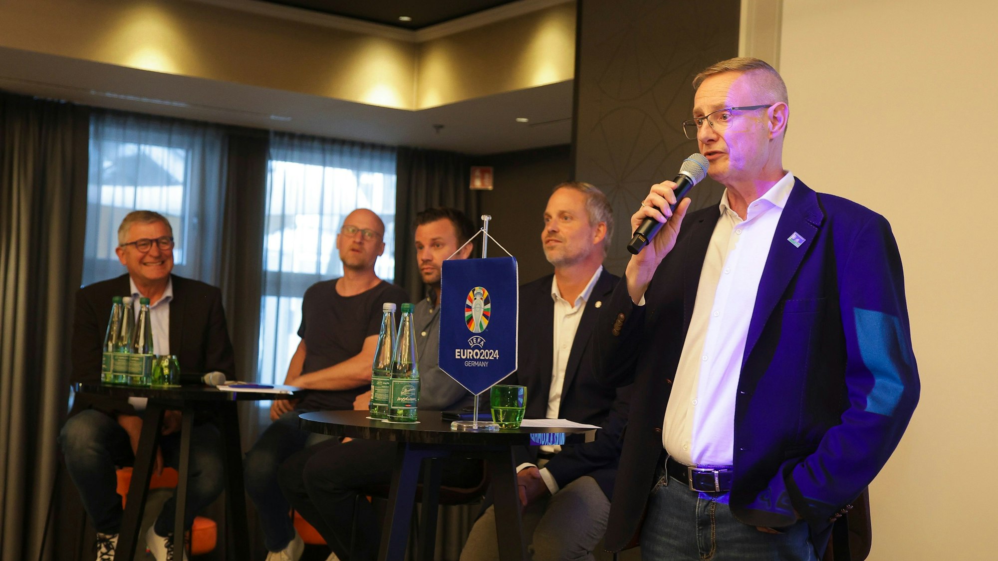 Fünf Männer auf einem Podium, einer spricht mit Mikrofon in der Hand.