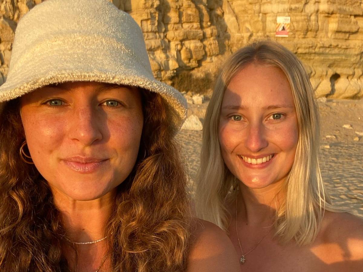 Zwei Frauen machen an einem Strand ein Selfie und grinsen in die Kamera.
