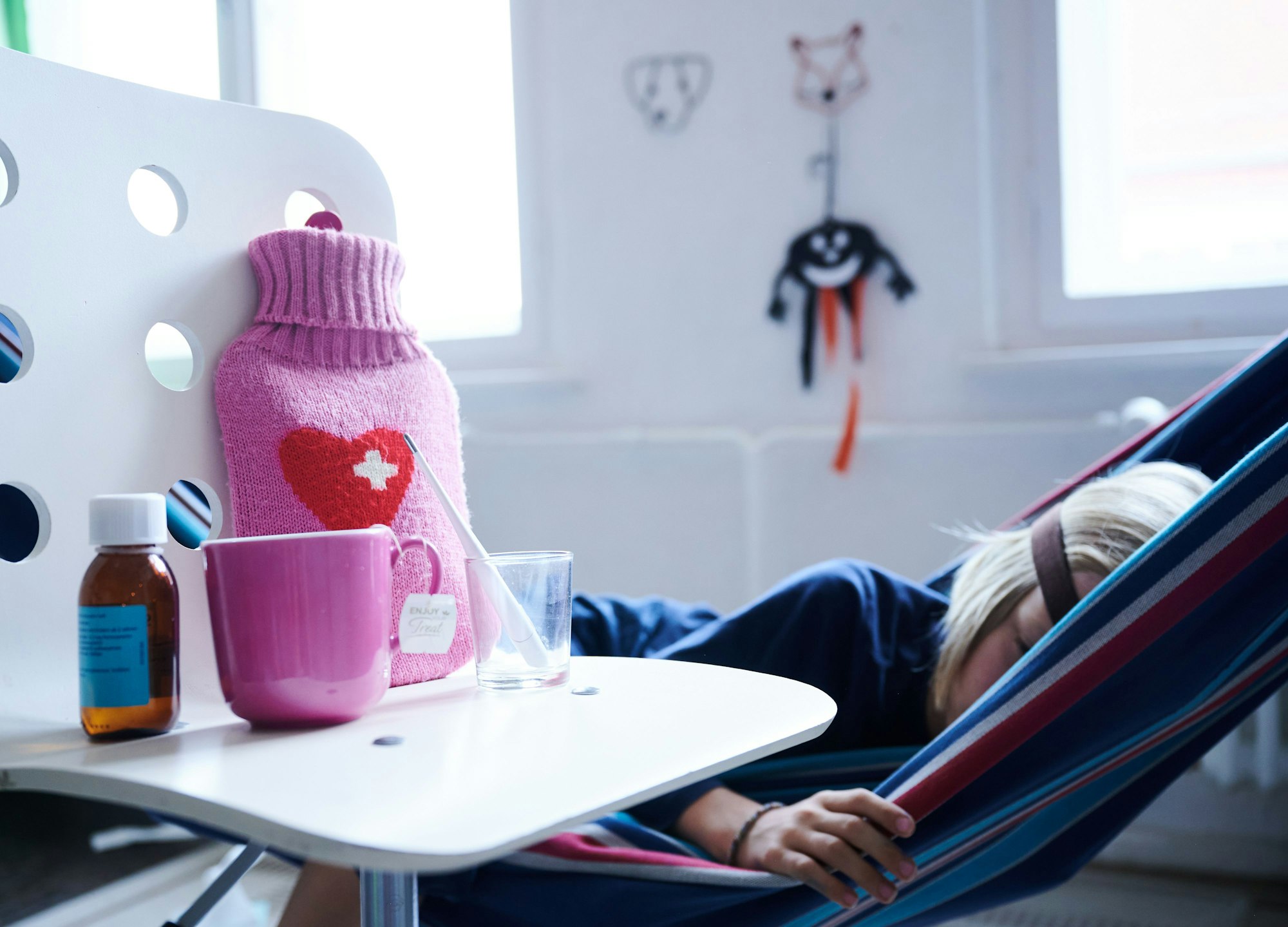 Fieberthermometer, Hustensaft, Wärmflasche und eine Tasse Tee stehen auf einem Stuhl, während ein Kind im Hintergrund in einer Hängematte liegt.