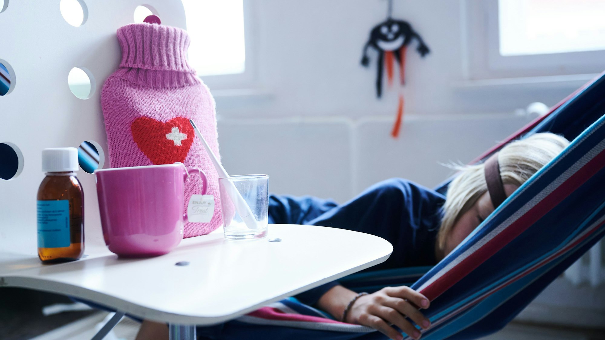 Foto eines kranken Kindes, das in einer Hängematte liegt. Neben ihm auf dem Tisch stehen Hustensaft, eine Tasse, ein Glas und eine Wärmflasche.