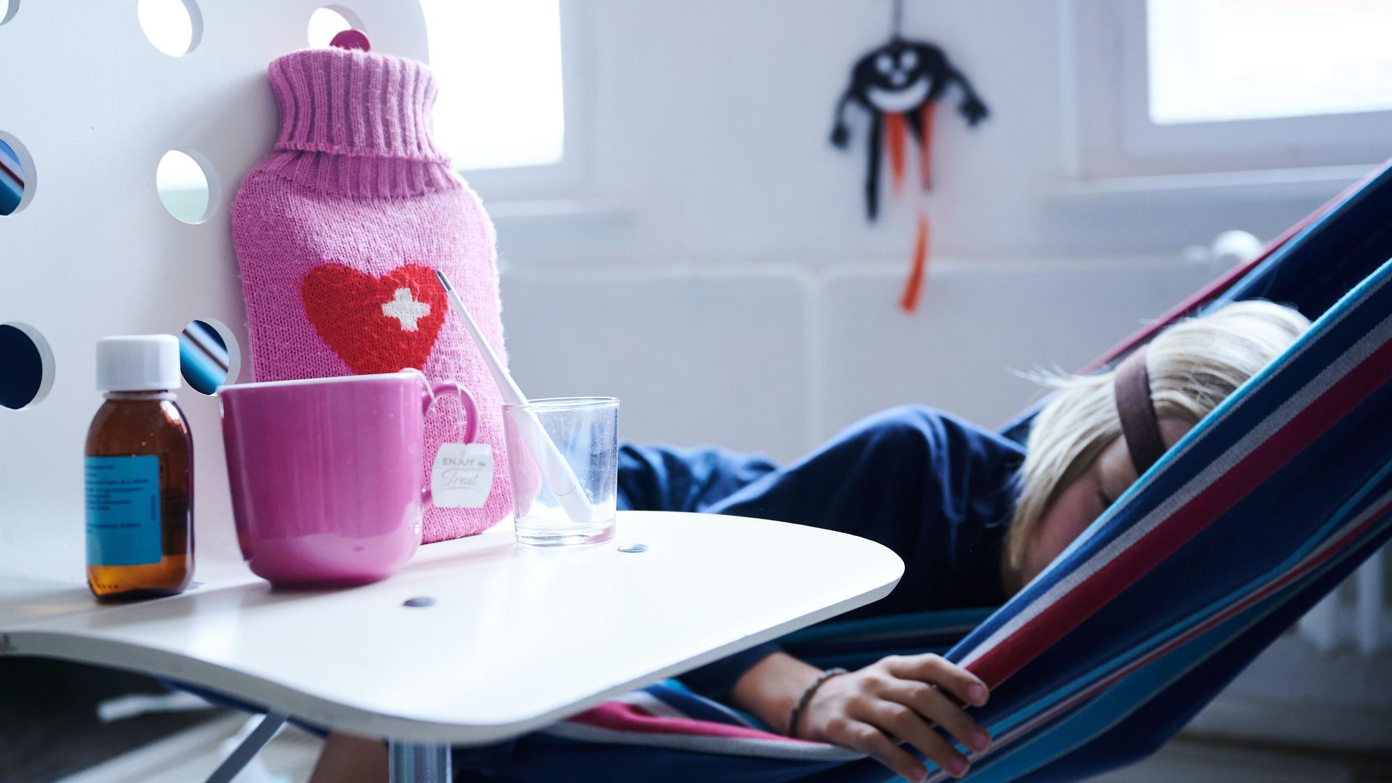Fieberthermometer, Hustensaft, Wärmflasche und eine Tasse Tee stehen auf einem Stuhl, während ein Kind im Hintergrund in einer Hängematte liegt.