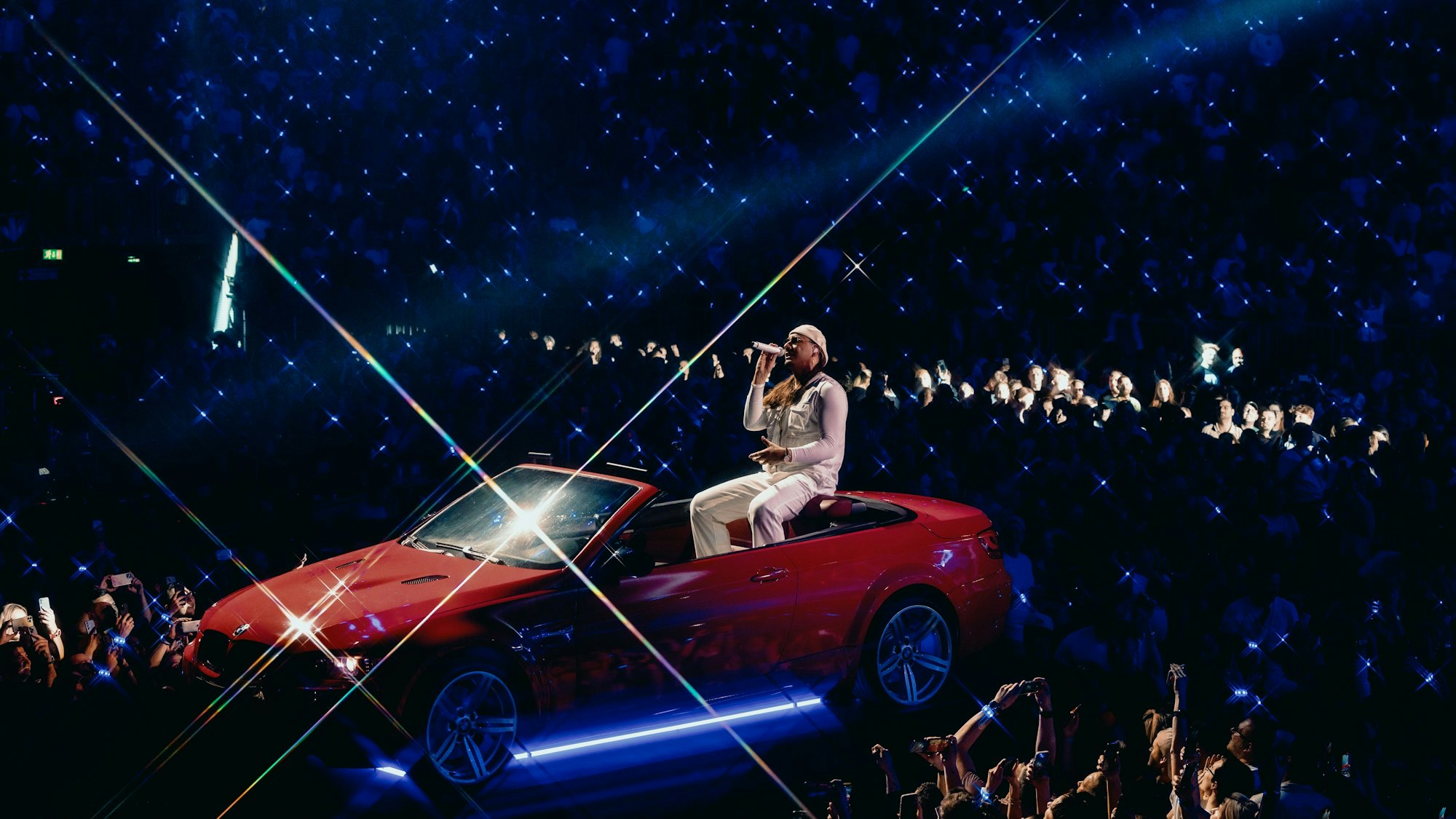 Apache 207 sitzt hinten auf einem roten Cabrio und singt.