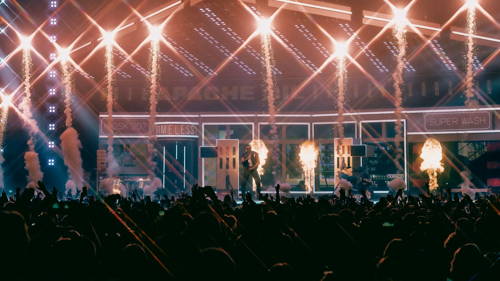 Apache 207 steht auf der Bühne in der Lanxess-Arena. Vor ihm sieht man die Arme der jubelnden Fans im Innenraum.
