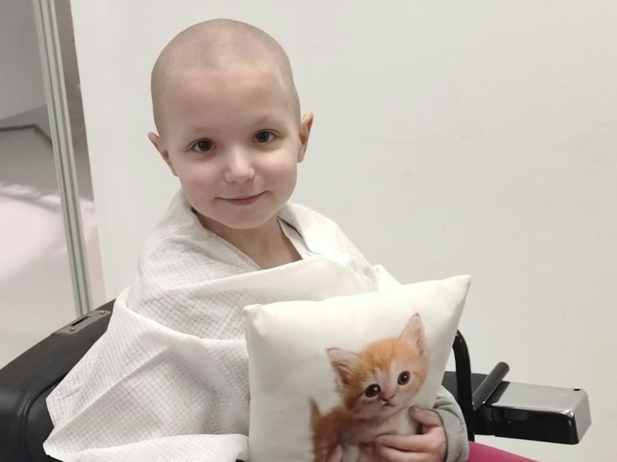 Ein kleines Mädchen ohne Haare hält ein Kissen mit einem Katzenbild vor sich.