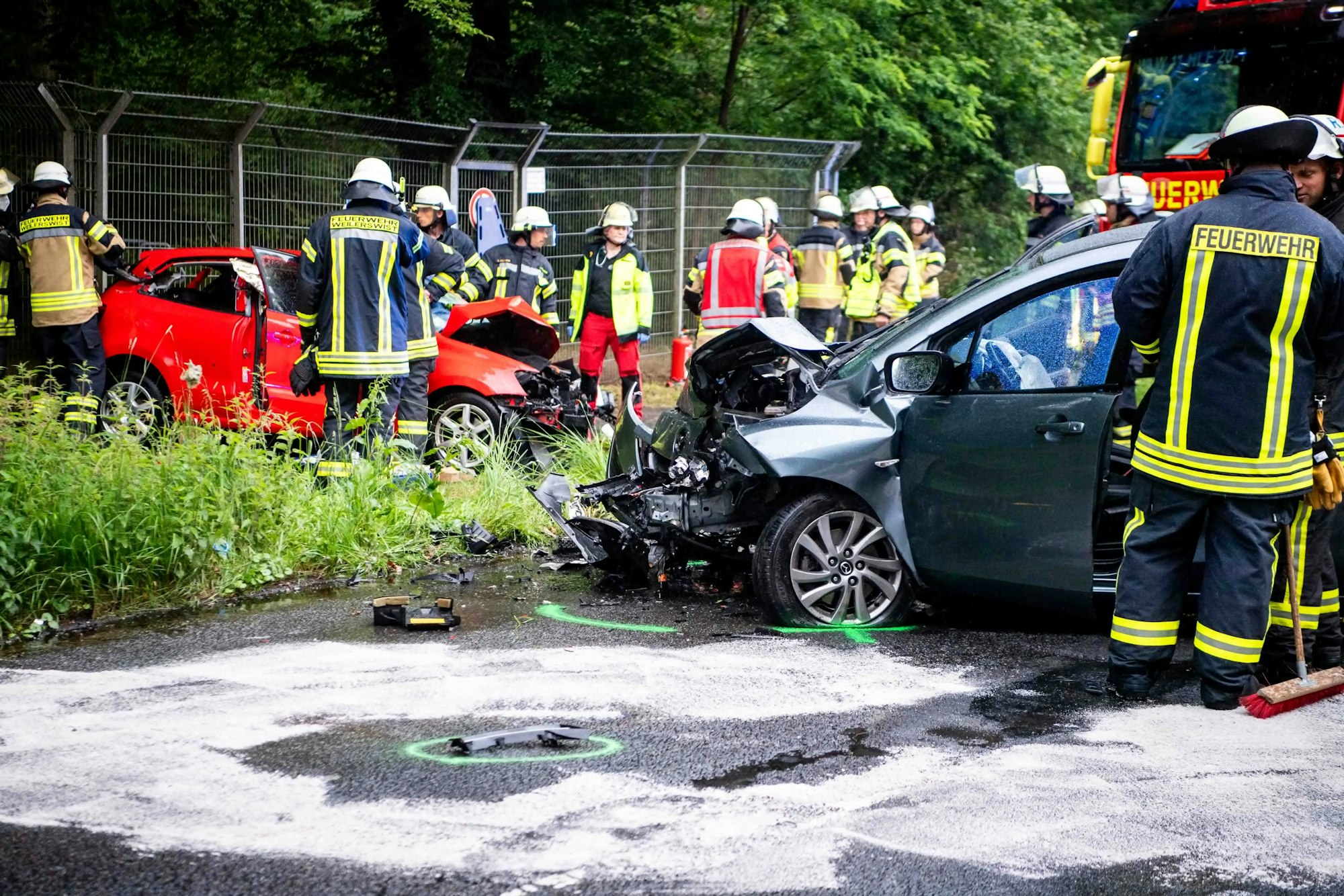 Einsatzkräfte der Feuerwehr stehen an zwei stark beschädigten Autos, von denen sich eines quer auf der Fahrbahn, das andere auf dem Gehweg befindet.