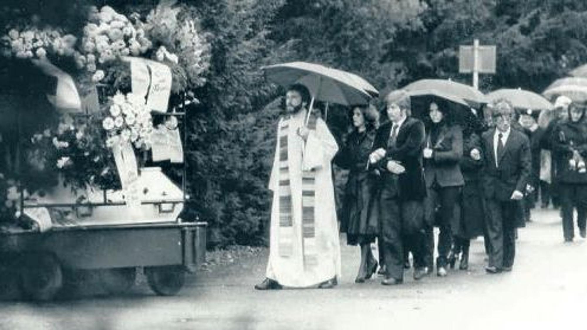 Ein Pfarrer und zahlreiche Trauernde folgen unter Regenschirmen einem Sarg.