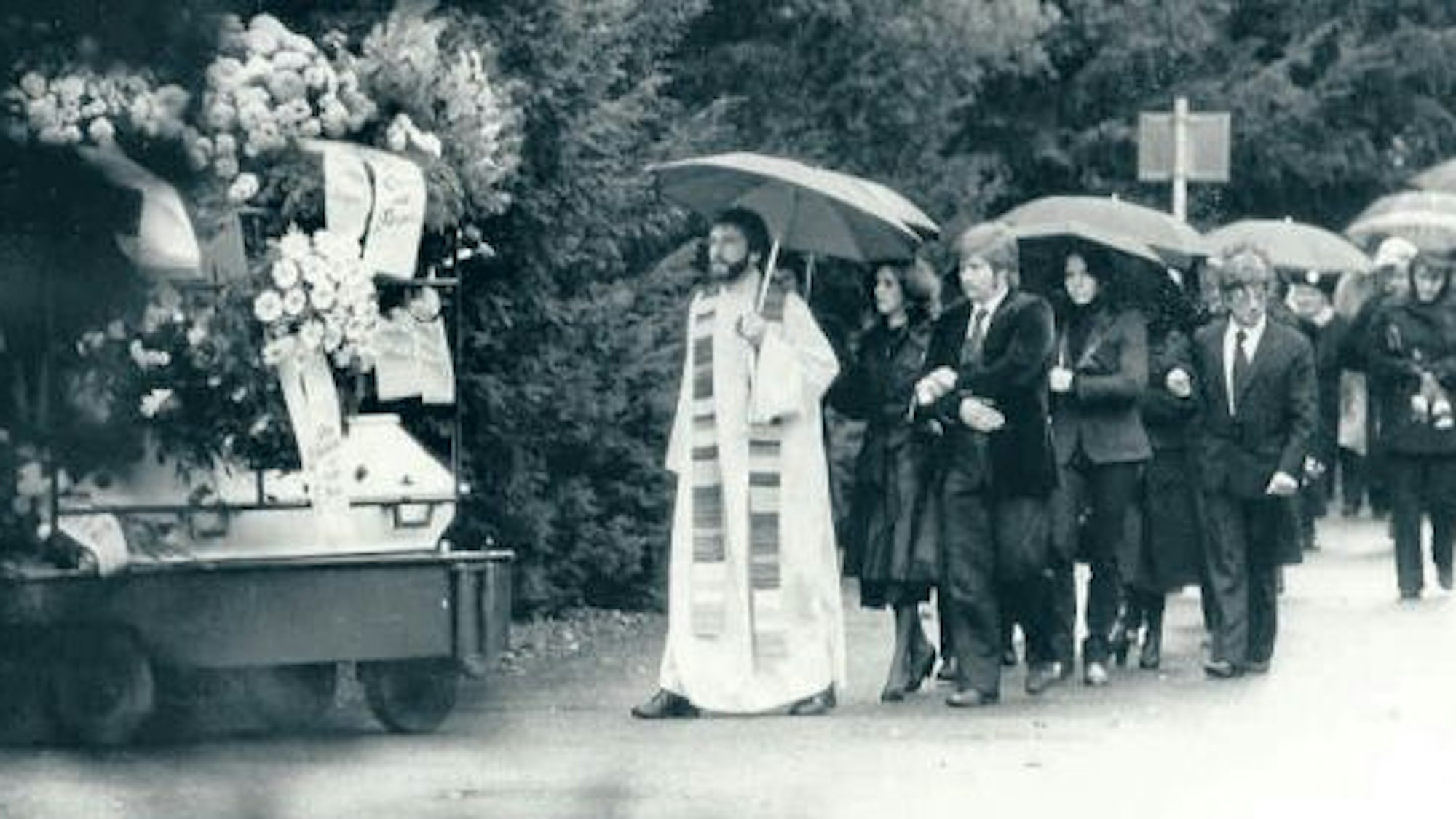Ein Pfarrer und zahlreiche Trauernde folgen unter Regenschirmen einem Sarg.