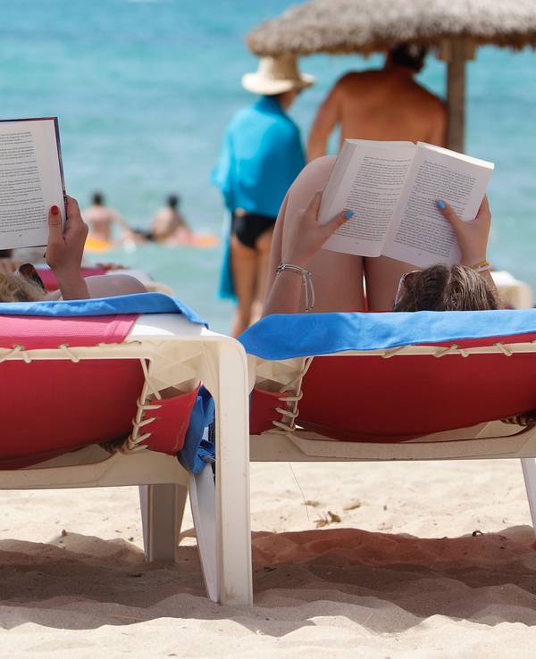 Zwei Frauen liegen am Strand „El Arenal“ auf Mallorca auf Liegestühlen und lesen in ihren Büchern, hier im August 2019.