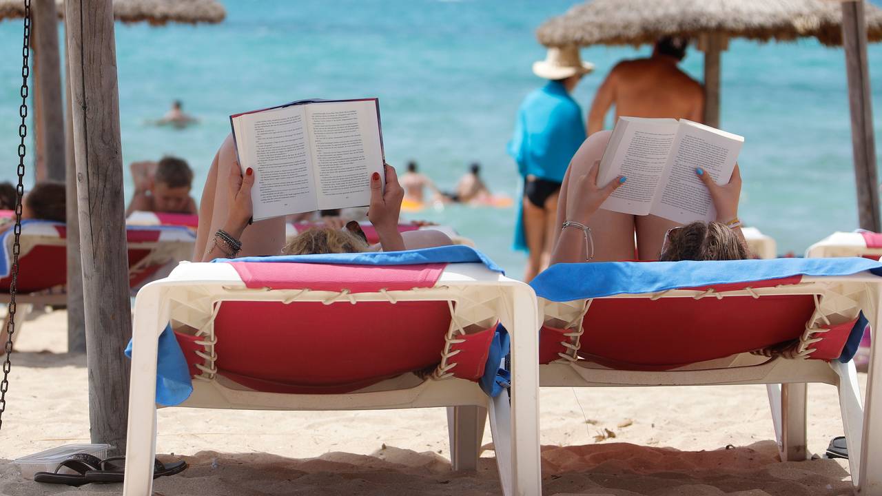 Zwei Frauen liegen am Strand „El Arenal“ auf Mallorca auf Liegestühlen und lesen in ihren Büchern, hier im August 2019.