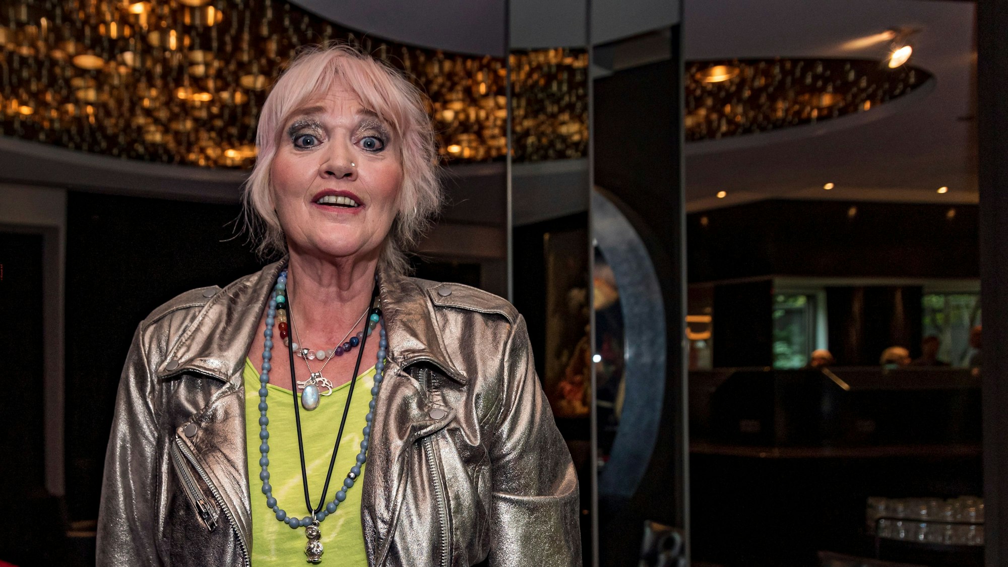 Gaby Köster steht in einer Lobby in Köln und schaut mir fröhlichem Gesicht in die Kamera.