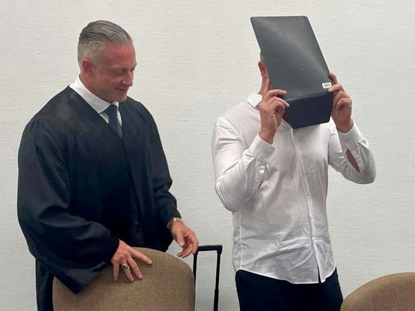 Angeklagter hält sich vor Gericht einen Aktenordner vor das Gesicht, daneben steht sein Verteidiger.