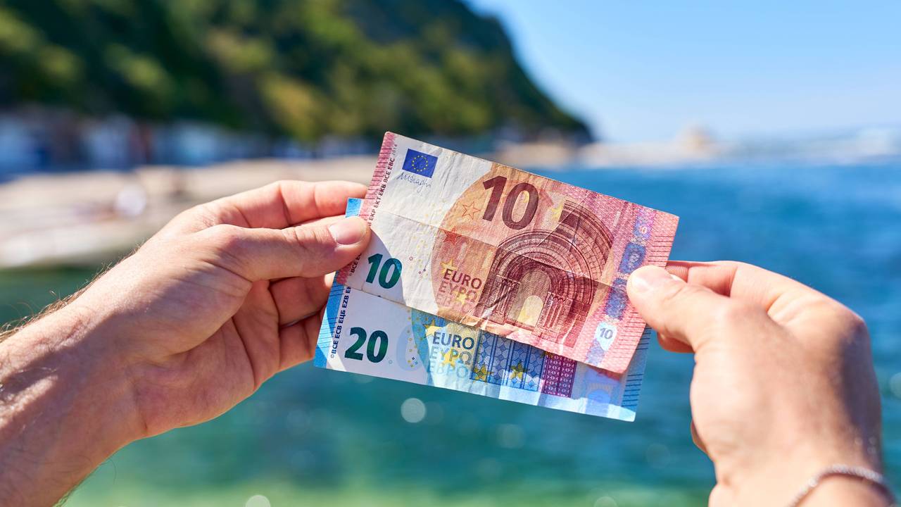 Dieses Symbolfoto zeigt: Ein Mann hält einen 20-Euro-Schein und einen 10-Euro-Schein an einem Strand in Italien in die Höhe.