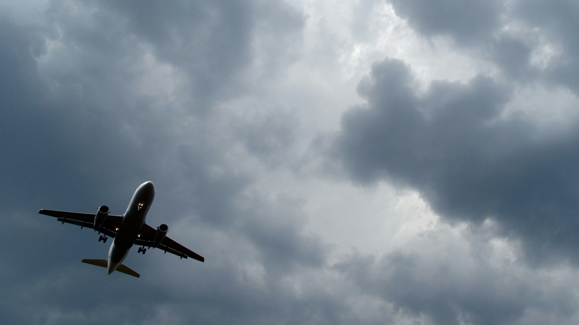 Ein Flugzeug am Himmel über Köln, im Hintergrund sind dunkle Wolken zu sehen, die ein Gewitter ankündigen.