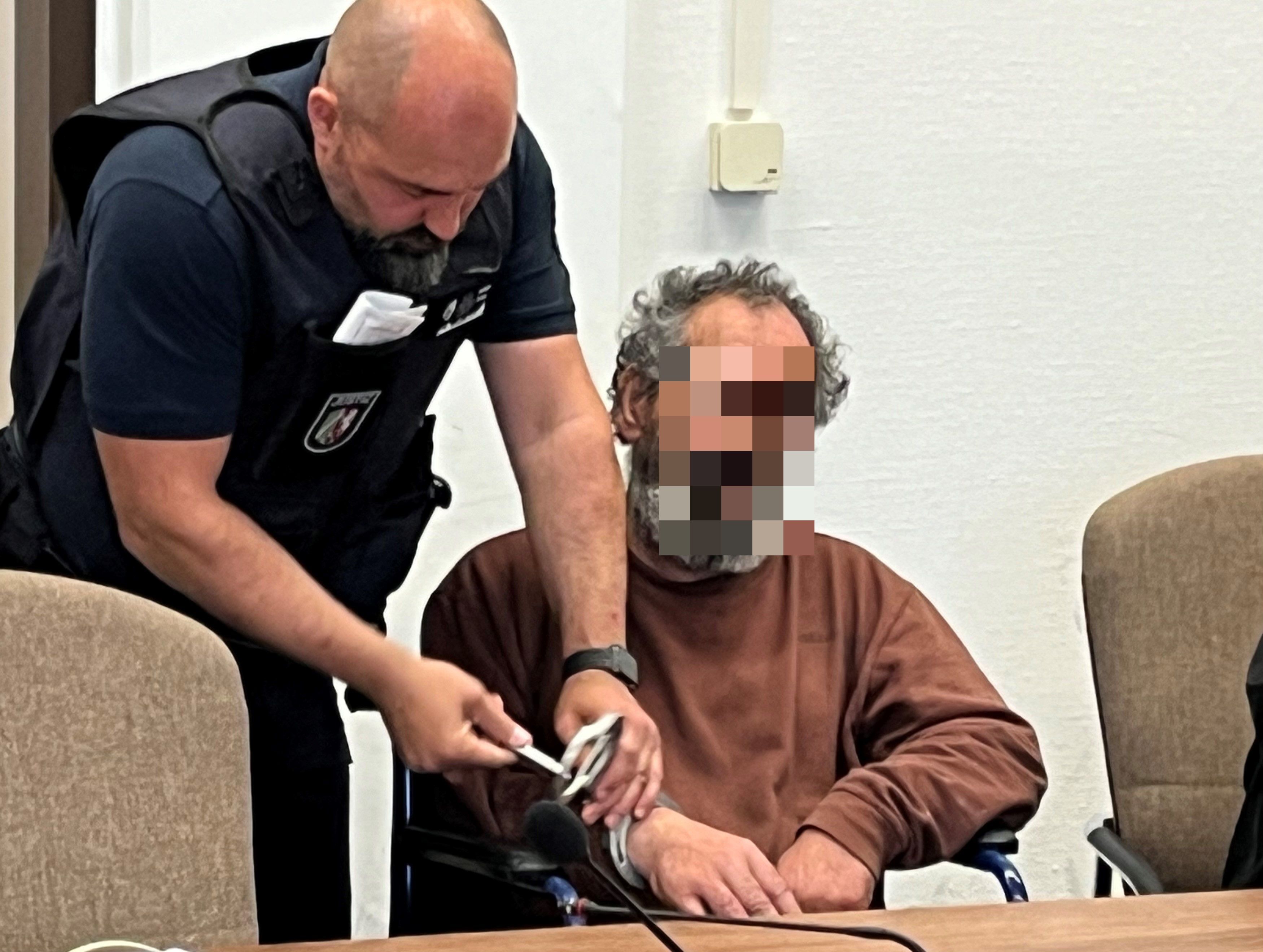 Ein Wachtmeister löst eine Handschelle, mit der ein Mann an seinen Rollstuhl gefesselt war.