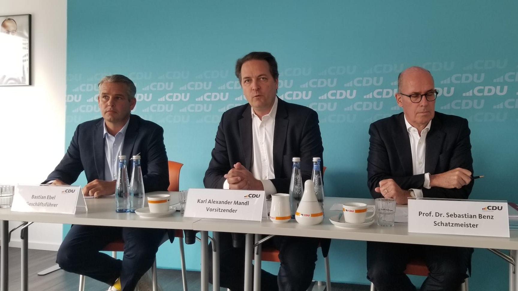 Drei Männer sitzen während einer Pressekonferenz vor einem großen Schreibtisch.