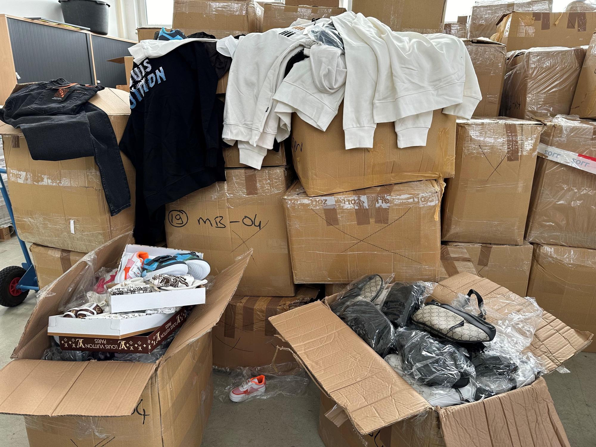 Ein Berg von Kartons, zwei davon sind geöffnet, sie sind voll mit Produktfälschungen, wie Taschen. Über anderen Kartons hängen Sweatshirts, auf einem steht „Louis Vuitton“.
