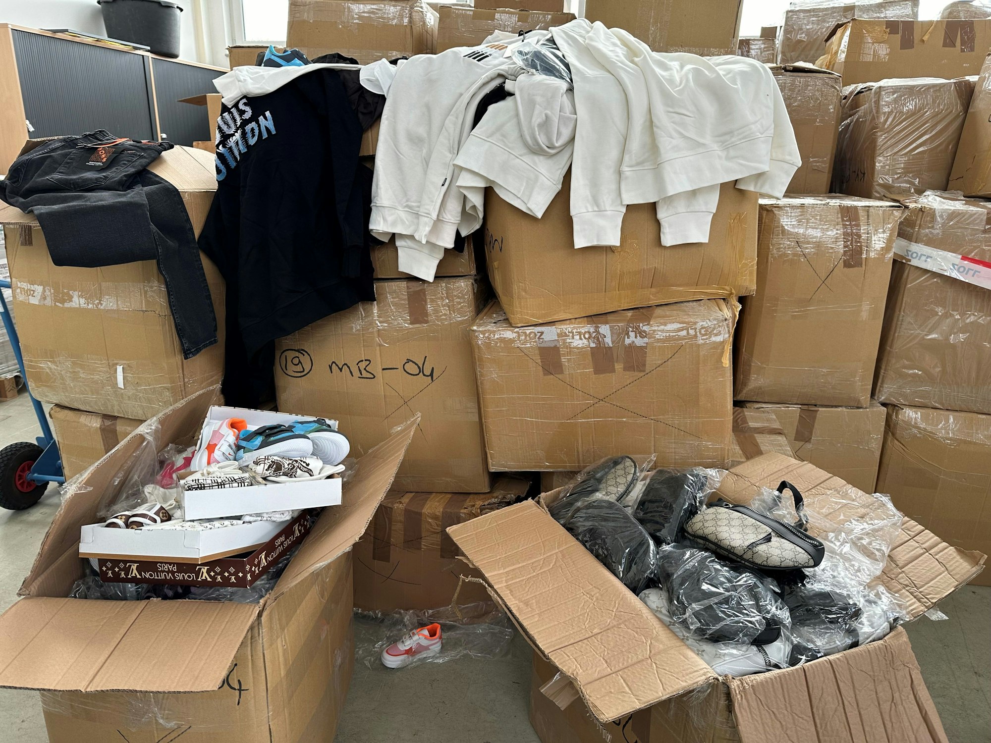 Ein Berg von Kartons, zwei davon sind geöffnet, sie sind voll mit Produktfälschungen, wie Taschen. Über anderen Kartons hängen Sweatshirts, auf einem steht „Louis Vuitton“.