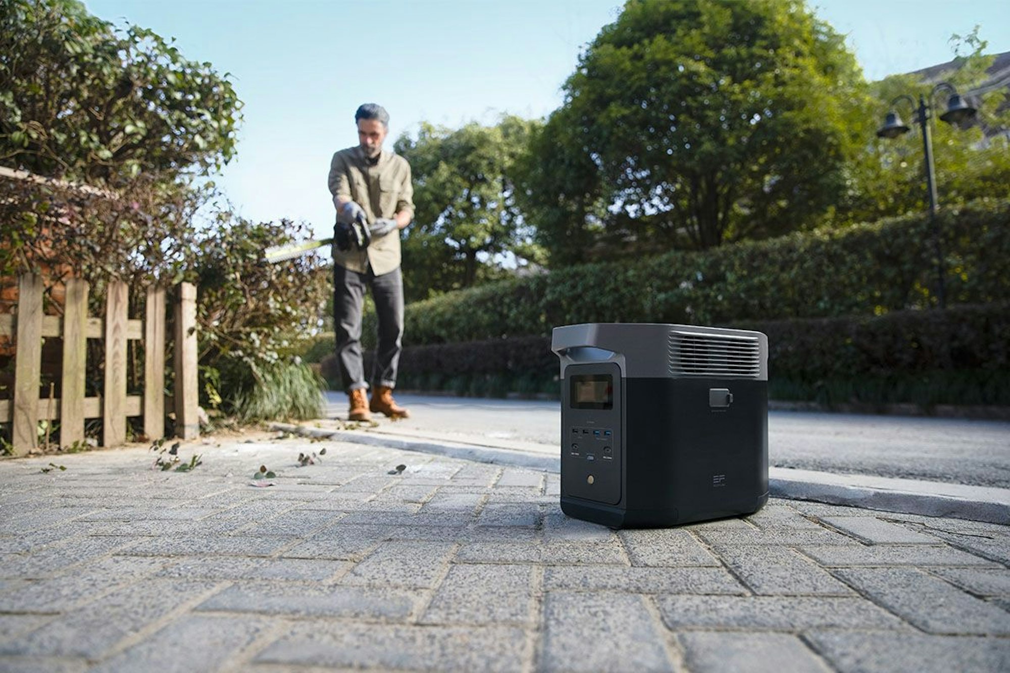 EcoFlow Powerstation steht auf dem Boden, ein Mann schneidet im Hintergrund die Hecke.