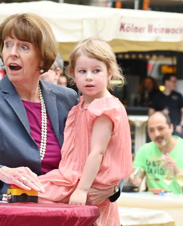 Oberbürgermeisterin Henriette Reker drückt den roten Knopf, der die Brunnenpumpe aktiviert. Dabei hat sie die Tochter von Jennifer Glashagen, Volt-Fraktionsvorsitzende, auf dem Arm.