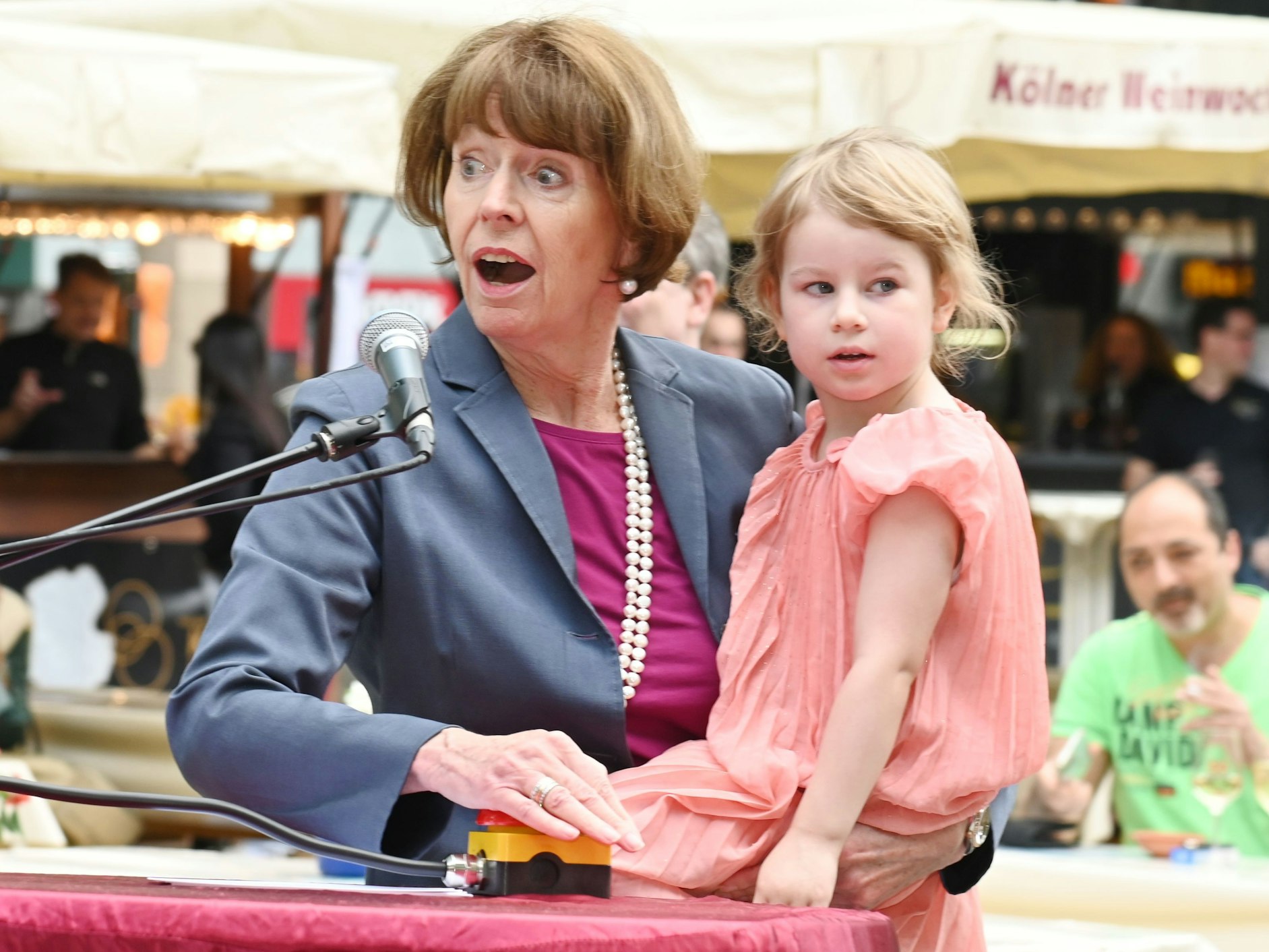 Oberbürgermeisterin Henriette Reker drückt den roten Knopf, der die Brunnenpumpe aktiviert. Dabei hat sie die Tochter von Jennifer Glashagen, Volt-Fraktionsvorsitzende, auf dem Arm.