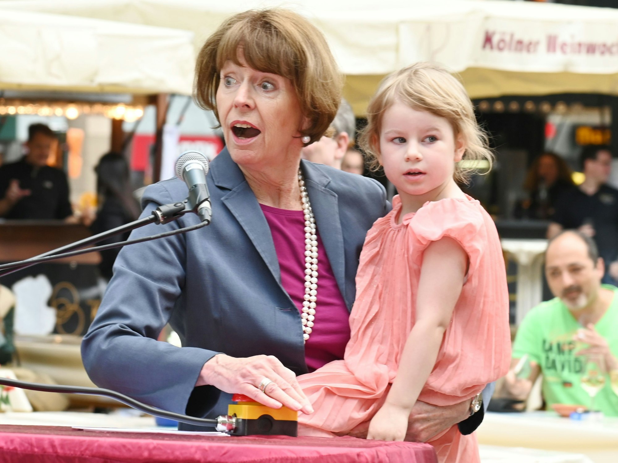 Oberbürgermeisterin Henriette Reker drückt den roten Knopf, der die Brunnenpumpe aktiviert. Dabei hat sie die Tochter von Jennifer Glashagen, Volt-Fraktionsvorsitzende, auf dem Arm.
